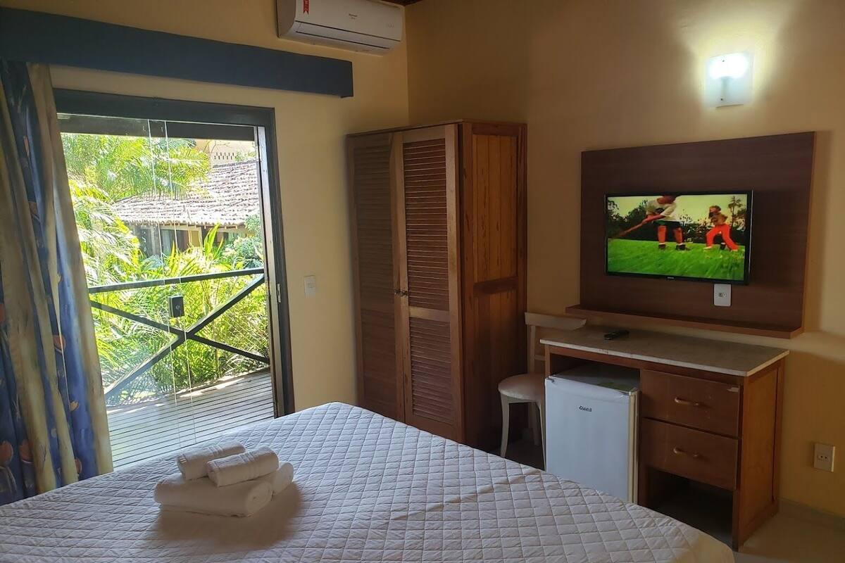 Habitación de hotel con cama, escritorio pequeño, televisión y balcón con vistas a la vegetación.