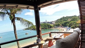 A wooden deck with a cushioned seating area, overlooking a beach with boats and a clear view of the sea. - Hotel La Foret & Beach Club (Búzios)
