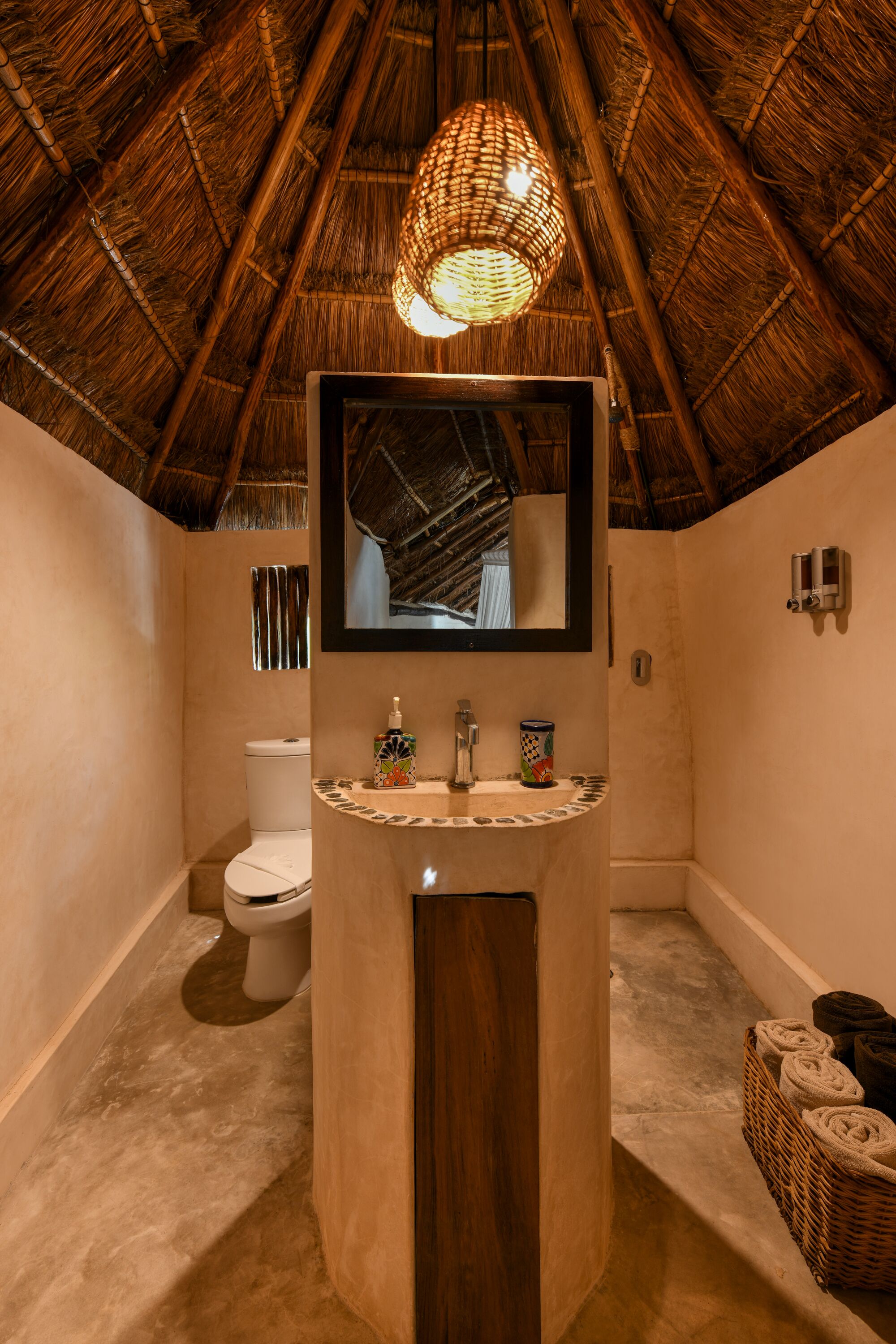 Een badkamer met rieten dak, een centraal geplaatst wastafelmeubel met spiegel, een toilet en een douchegedeelte.