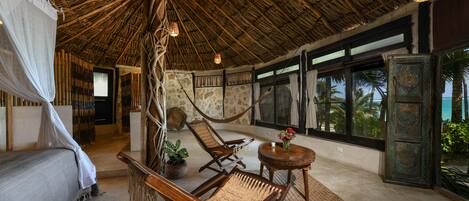 A room with a thatched roof, a bed with a mosquito net, two wicker chairs, a small table with a vase, and a view of palm trees through large windows.