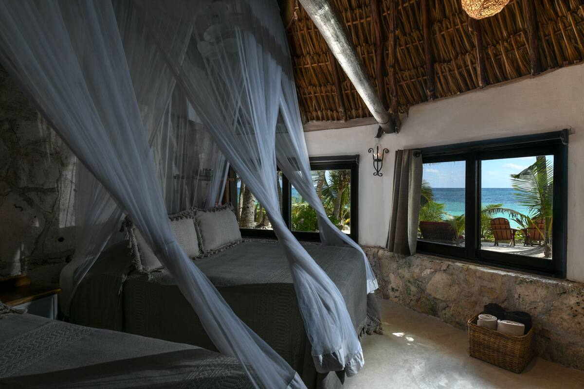 A bedroom with a bed under a canopy, a stone wall, a window with a view of the sea, and a wicker basket on the floor.