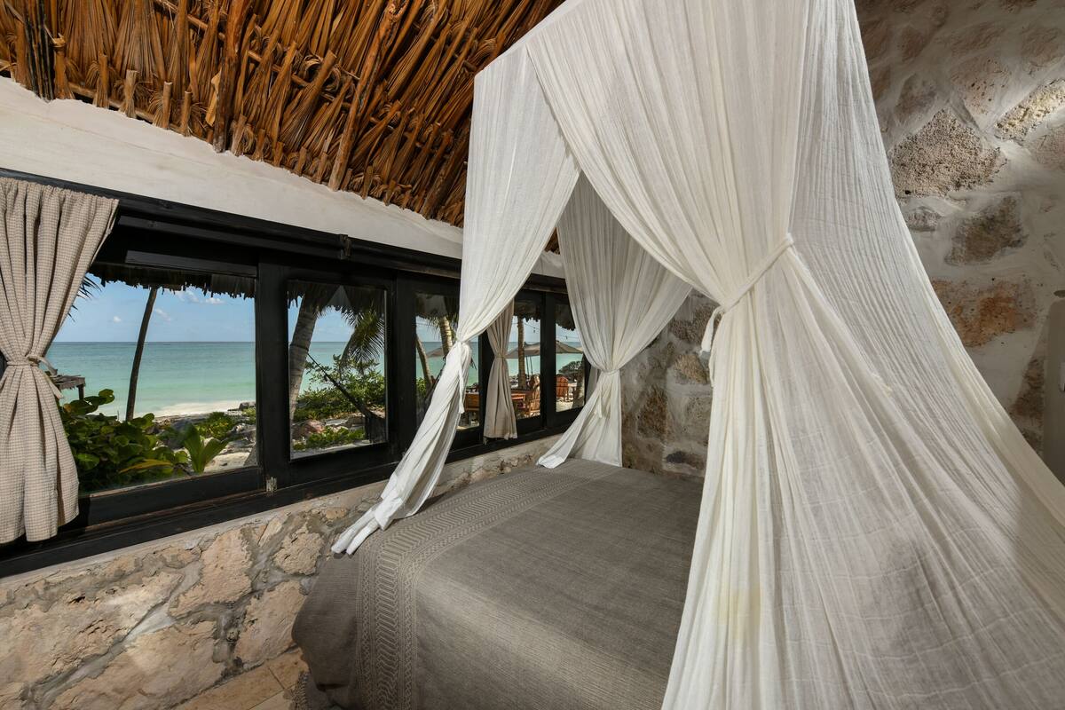 A room with a stone wall, a bed with a canopy, and a view of the beach and ocean through large windows.