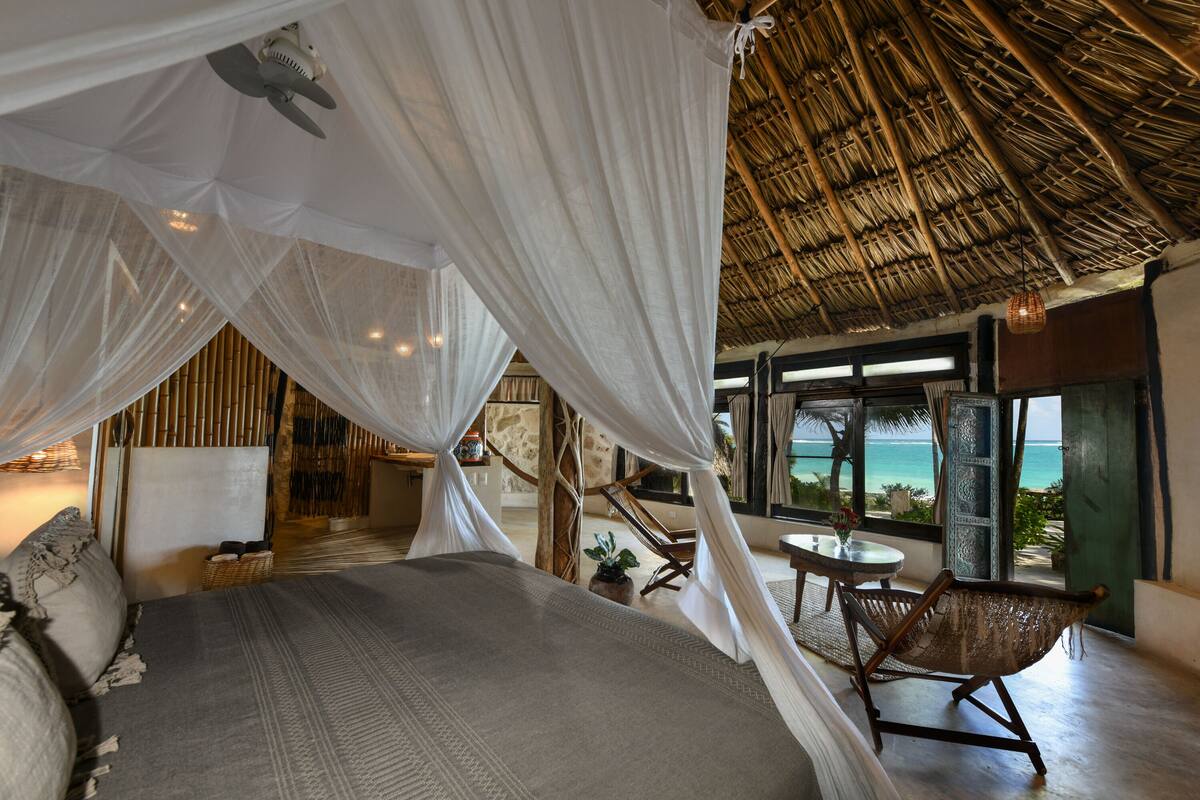 A bedroom with a canopy bed, a view of the ocean, and a thatched roof.
