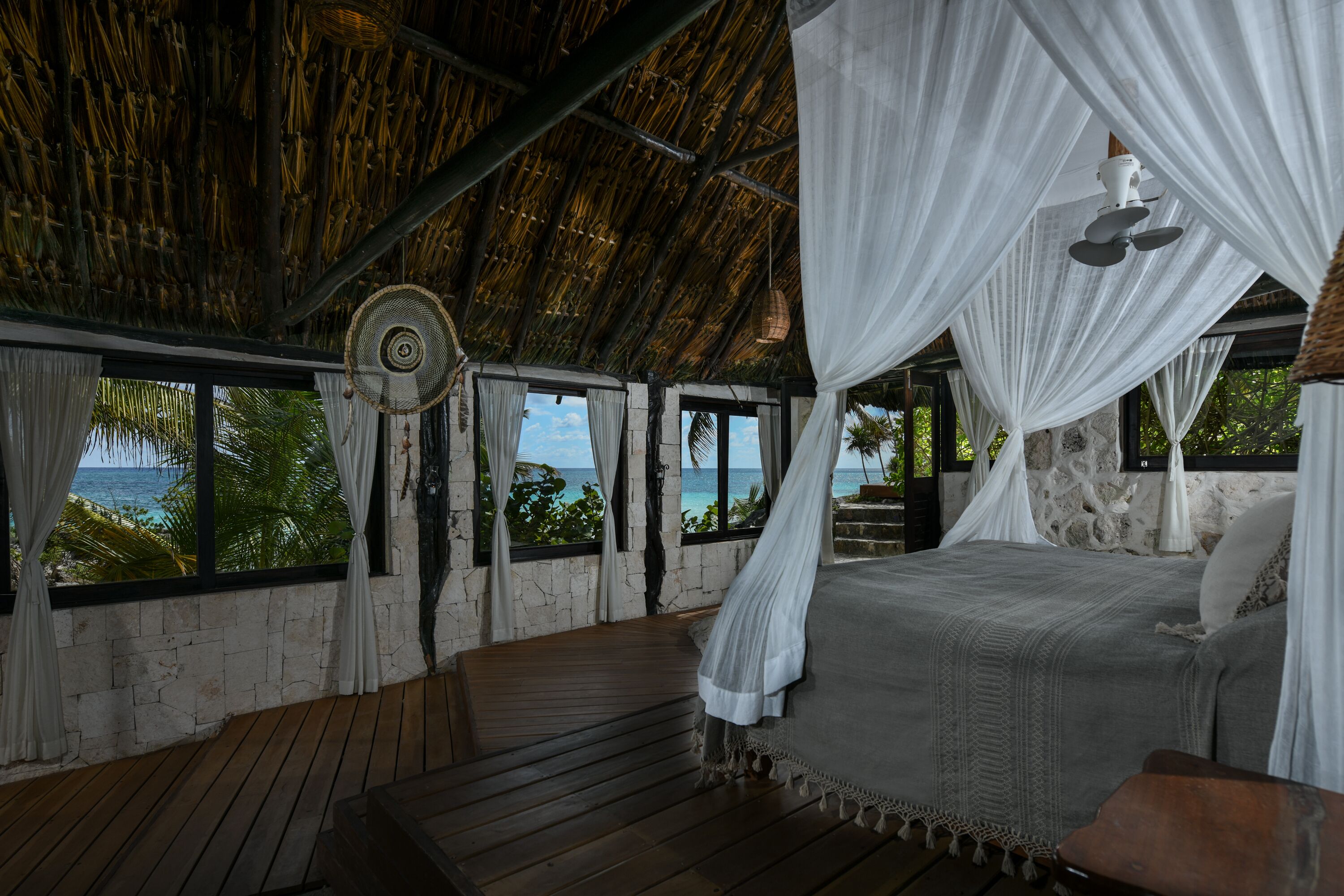 Una habitación con cama, piso de madera, techo de paja y ventanas con vistas a la playa.