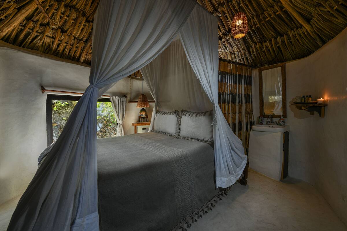 A bedroom with a canopy bed, a bedside table with a lamp, a mirror, and a window with a view of greenery.