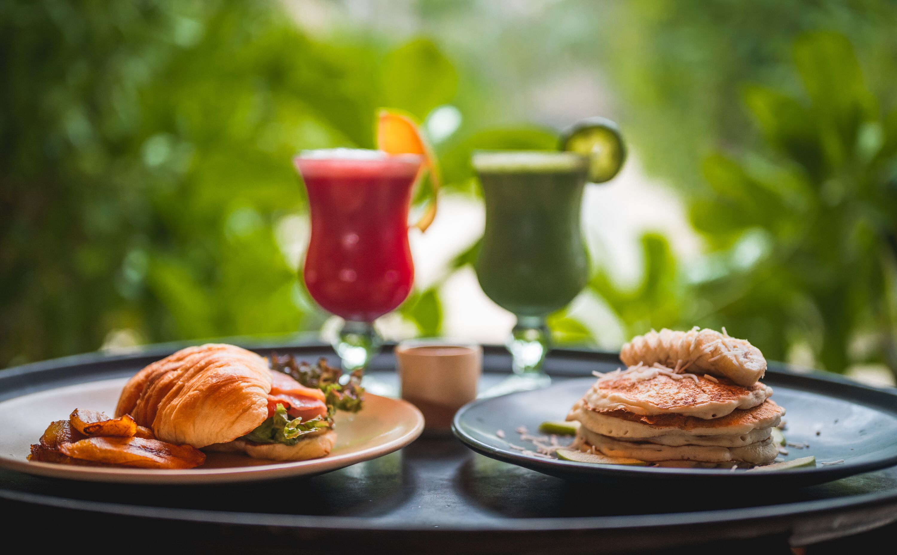 A breakfast setting with a croissant sandwich, bacon, pancakes, and two drinks on plates.