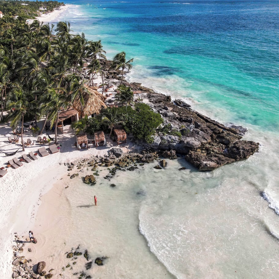 An aerial view of a beach resort with thatched-roof huts, lounge chairs, and a rocky shoreline.