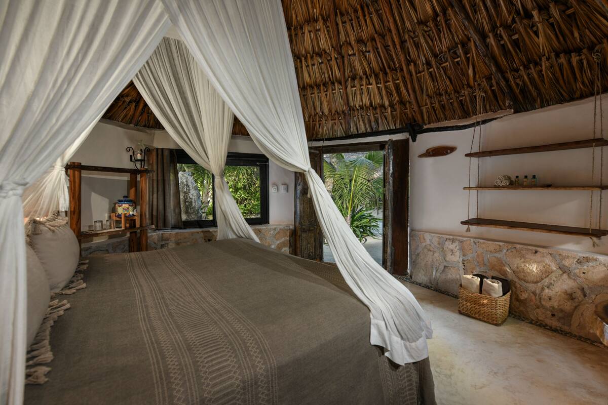 A room with a bed under a thatched roof, a stone wall, and a view of greenery outside.