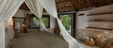 Habitación con cama bajo un tejado de paja, pared de piedra y vistas a la vegetación.