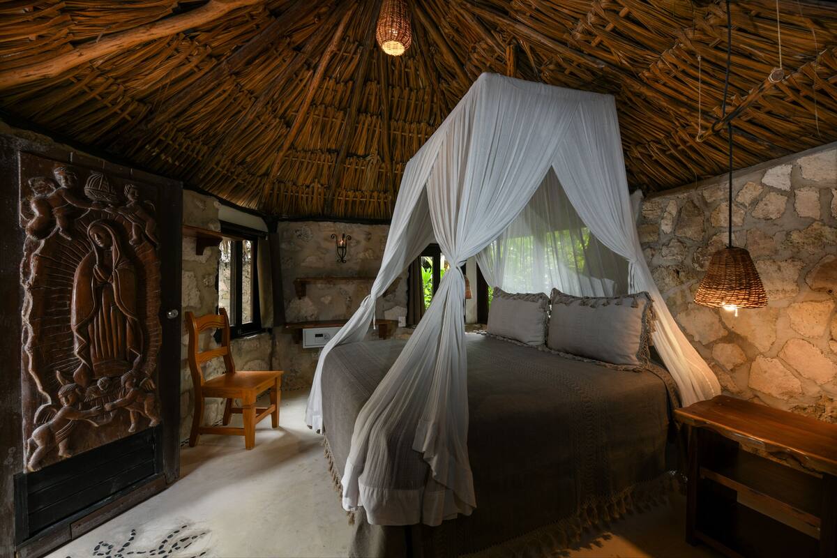 A room with a bed under a thatched roof, a stone wall, and a carved wooden panel.
