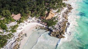 Vue aérienne d’un complexe hôtelier en bord de mer, avec une plage de sable, des huttes aux toits de chaume et une végétation luxuriante.