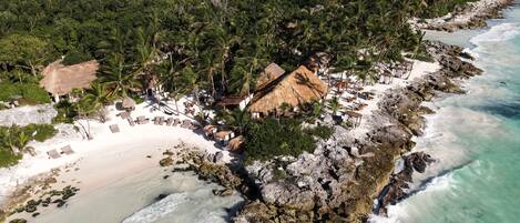 An aerial view of a beach resort with thatched-roof huts, white sandy beaches, and lush greenery.
