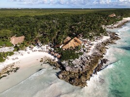 Vista aérea de un complejo turístico en la playa con cabañas de techo de paja, arena blanca y exuberante vegetación.