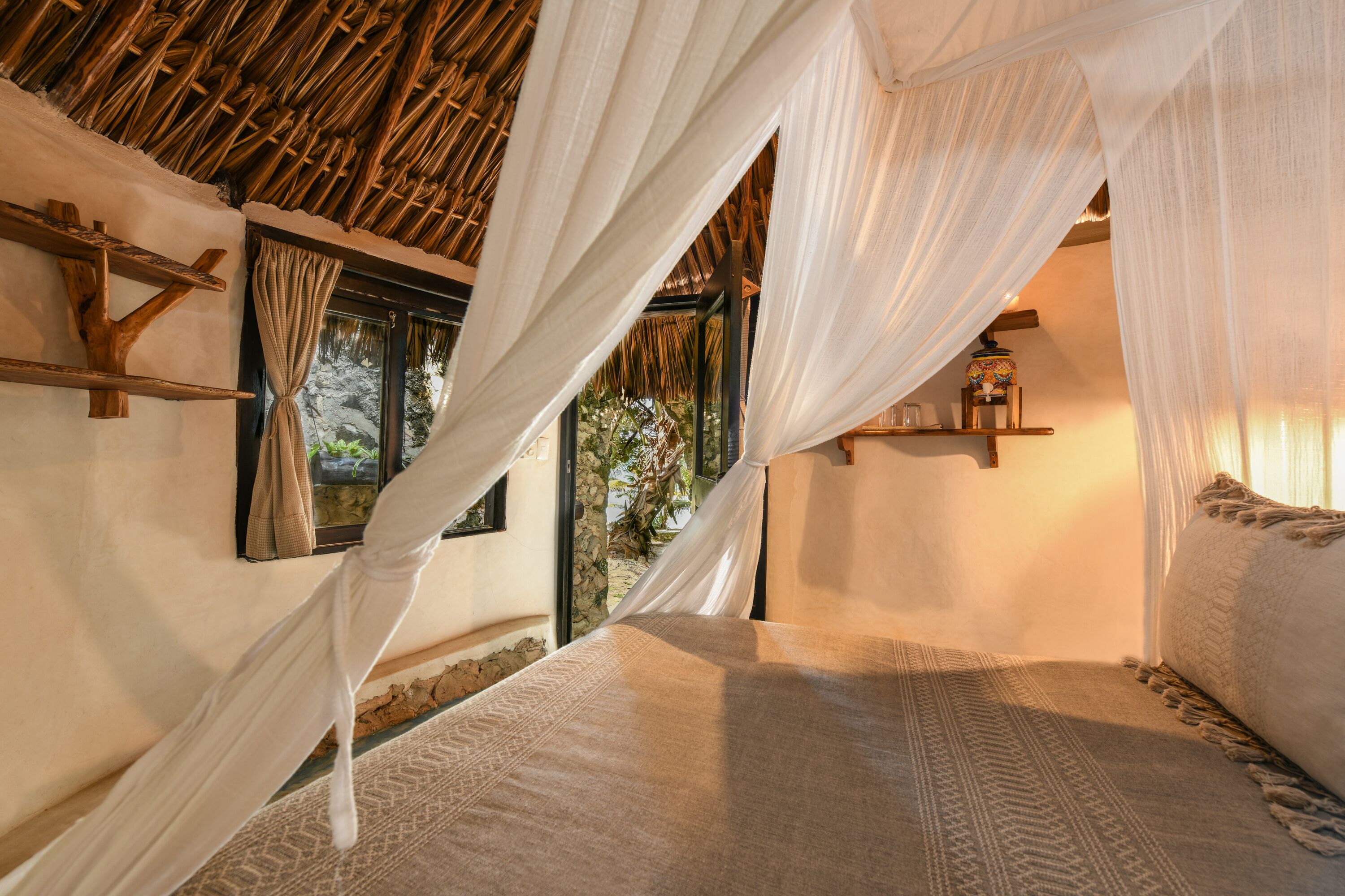 A room with a bed, a window with curtains, a wooden shelf, and a thatched roof.