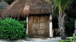 Une hutte traditionnelle à toit de chaume, dotée d’une porte en bois, entourée d’une végétation luxuriante et d’un palmier.