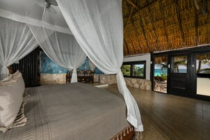 Une chambre spacieuse avec un grand lit, un baldaquin et une vue sur l’océan par une porte vitrée.