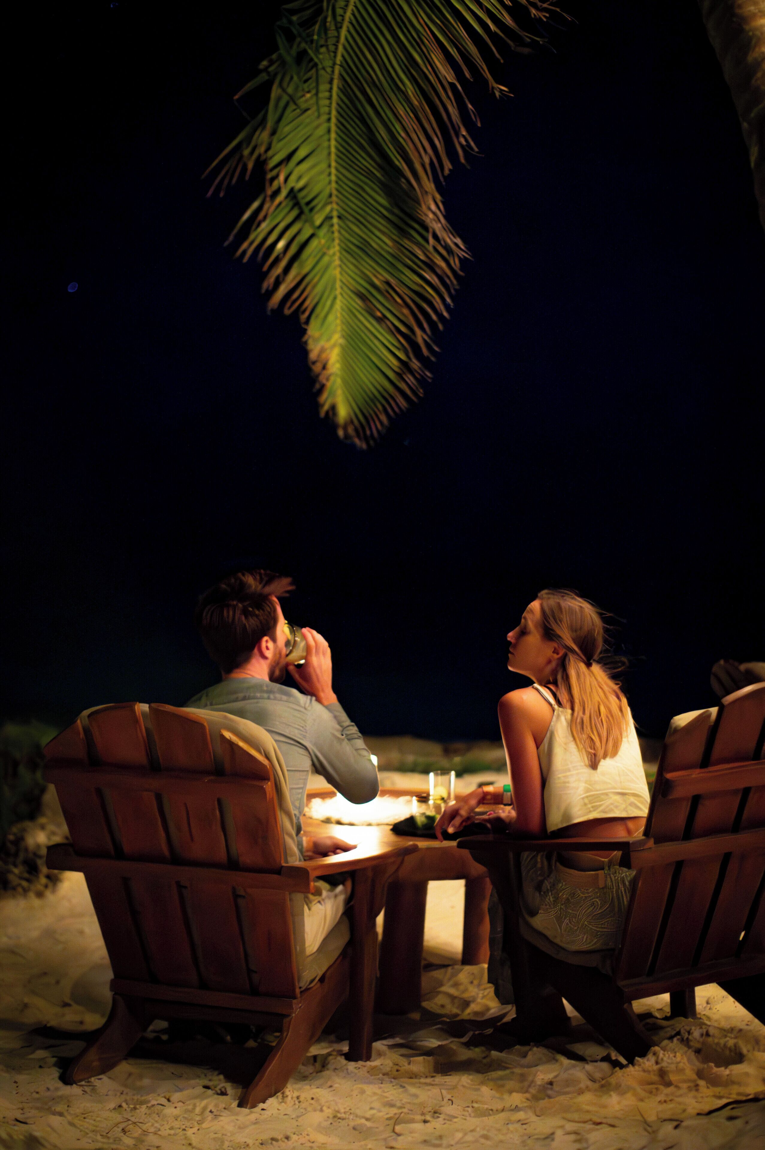 Two people sitting at a wooden table on a sandy beach at night, with a palm tree overhead.