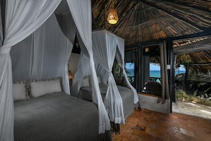 Una habitación con dos camas cubiertas por dos doseles blancos, techo de paja y vista al mar.