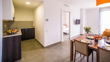 Una cocina moderna con un frutero, un comedor con una mesa dispuesta para dos personas, y un dormitorio visible a través de una puerta abierta.