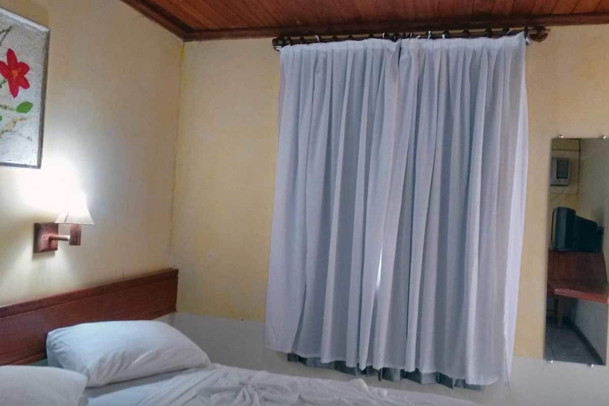 Ein Zimmer mit einem Bett, einer Wandlampe und einem großen Fenster mit weißen Vorhängen.