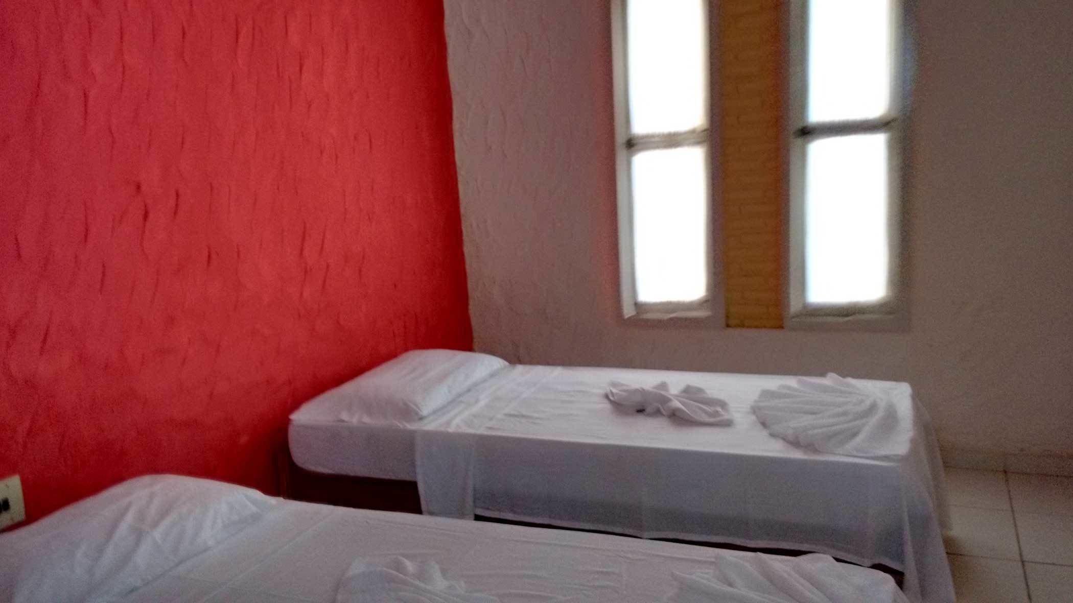 A room with two beds, one with a red wall and the other with a white wall. There is a window with a wooden blind.