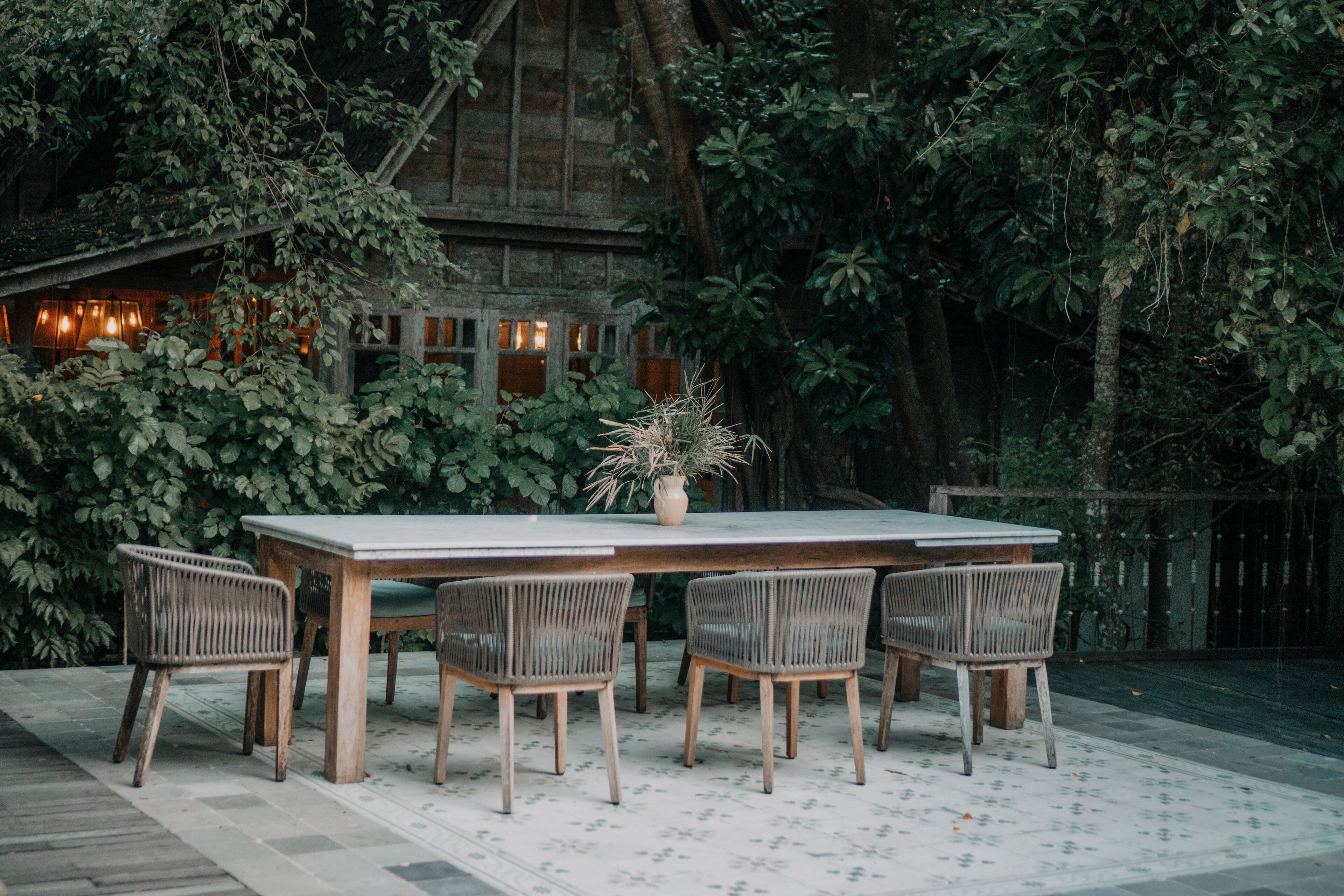 An outdoor dining area with a wooden table and wicker chairs, surrounded by lush greenery and a rustic building in the background.