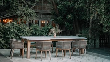 An outdoor dining area with a wooden table and wicker chairs, surrounded by lush greenery and a rustic building in the background.