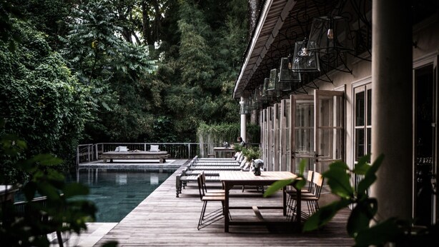 A wooden deck with tables and chairs overlooking a pool, surrounded by lush greenery. - BALQUISSE Heritage Hotel (Jimbaran)