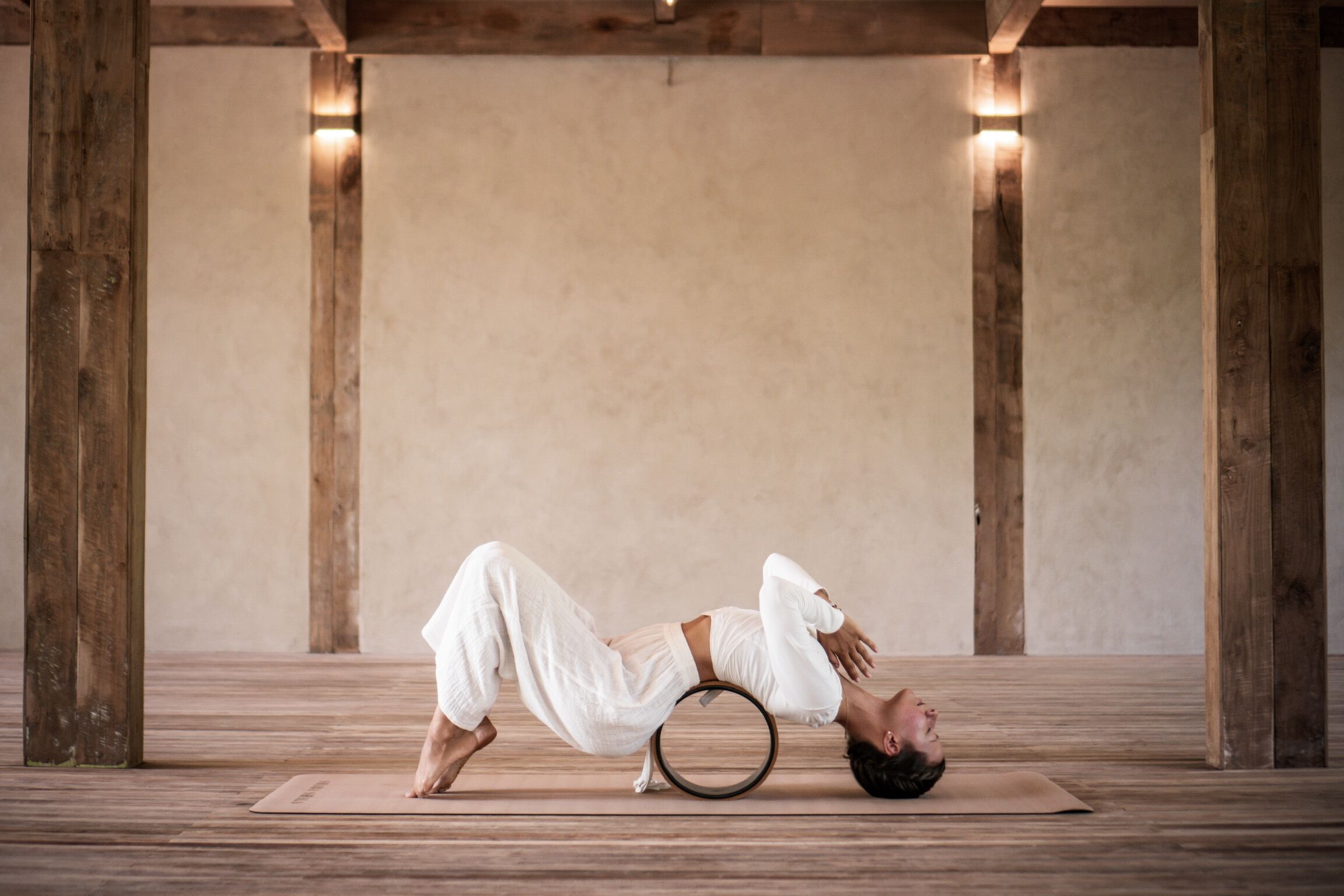 Una persona realizando una postura de yoga sobre una esterilla en una habitación con vigas de madera y pared lisa.
