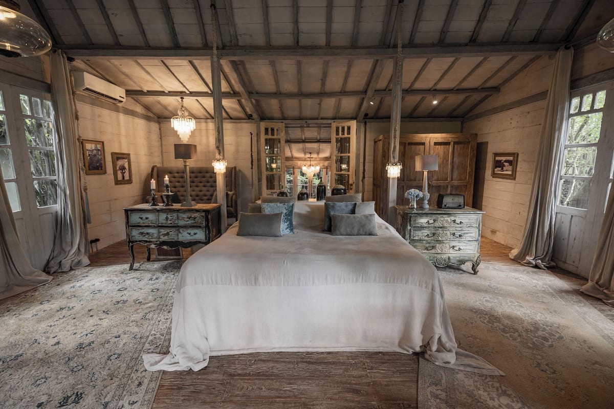 A spacious bedroom with a large bed, wooden ceiling beams, and a view through a doorway to a conservatory.
