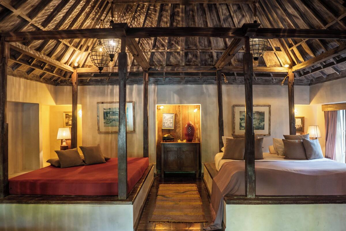 A room with a wooden ceiling, two beds with red and white bedding, a wooden nightstand with a lamp, and framed artwork on the walls.