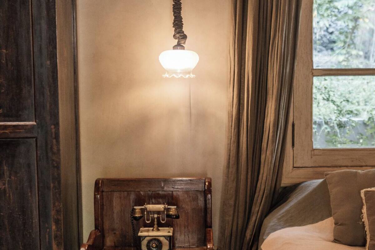 A room with a wooden nightstand holding a vintage telephone, a hanging light fixture, a bed with a beige blanket, and a window with curtains.