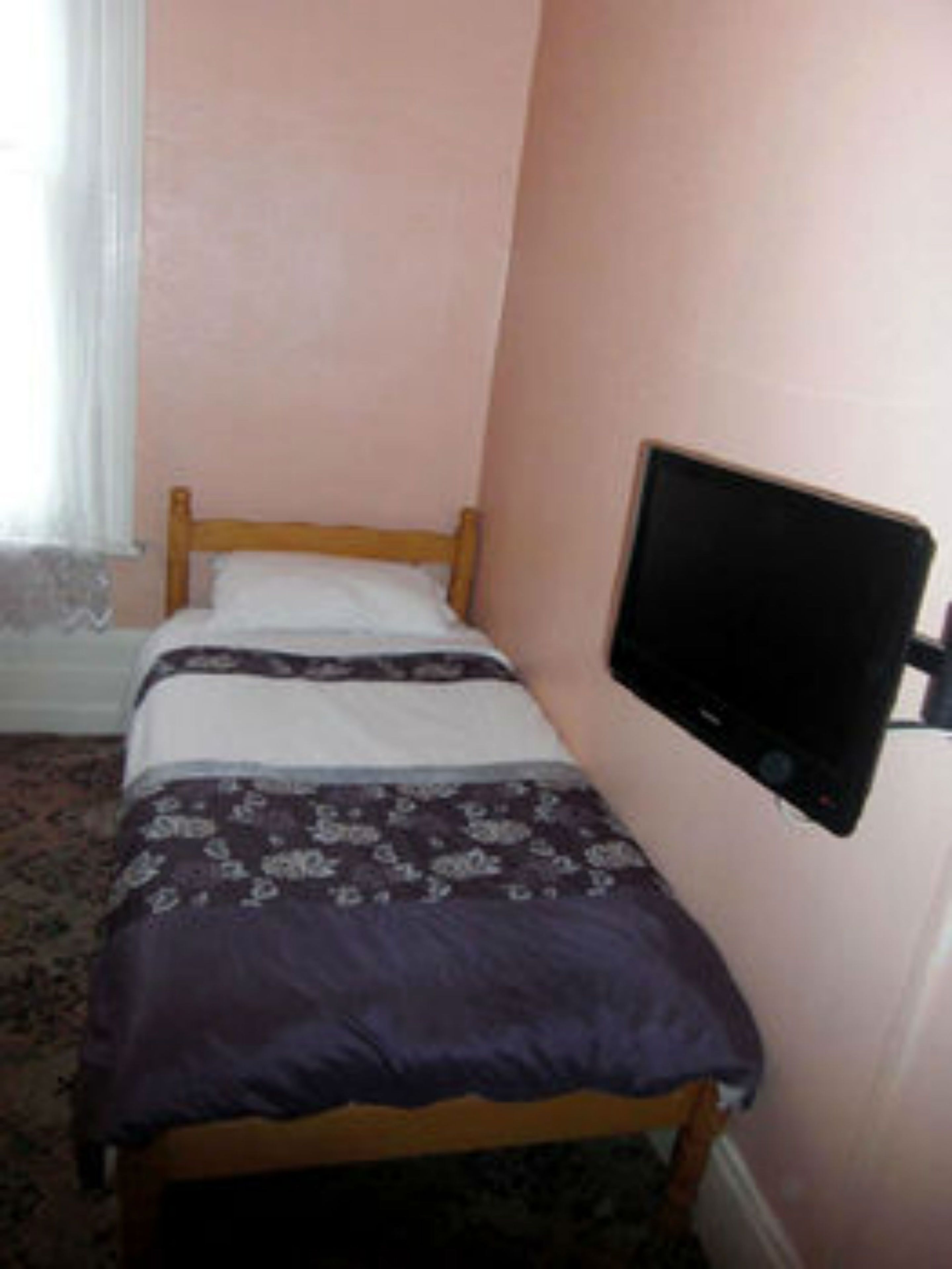 Habitació petita amb un llit individual, una televisió penjada a la paret i una finestra amb cortines.