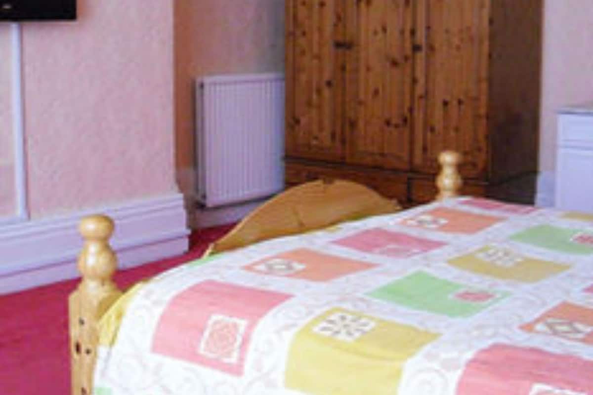 Een slaapkamer met een houten bedframe, een gewatteerde spreideken, een houten garderobe, een witte nachtkastje en een aan de muur bevestigde televisie.