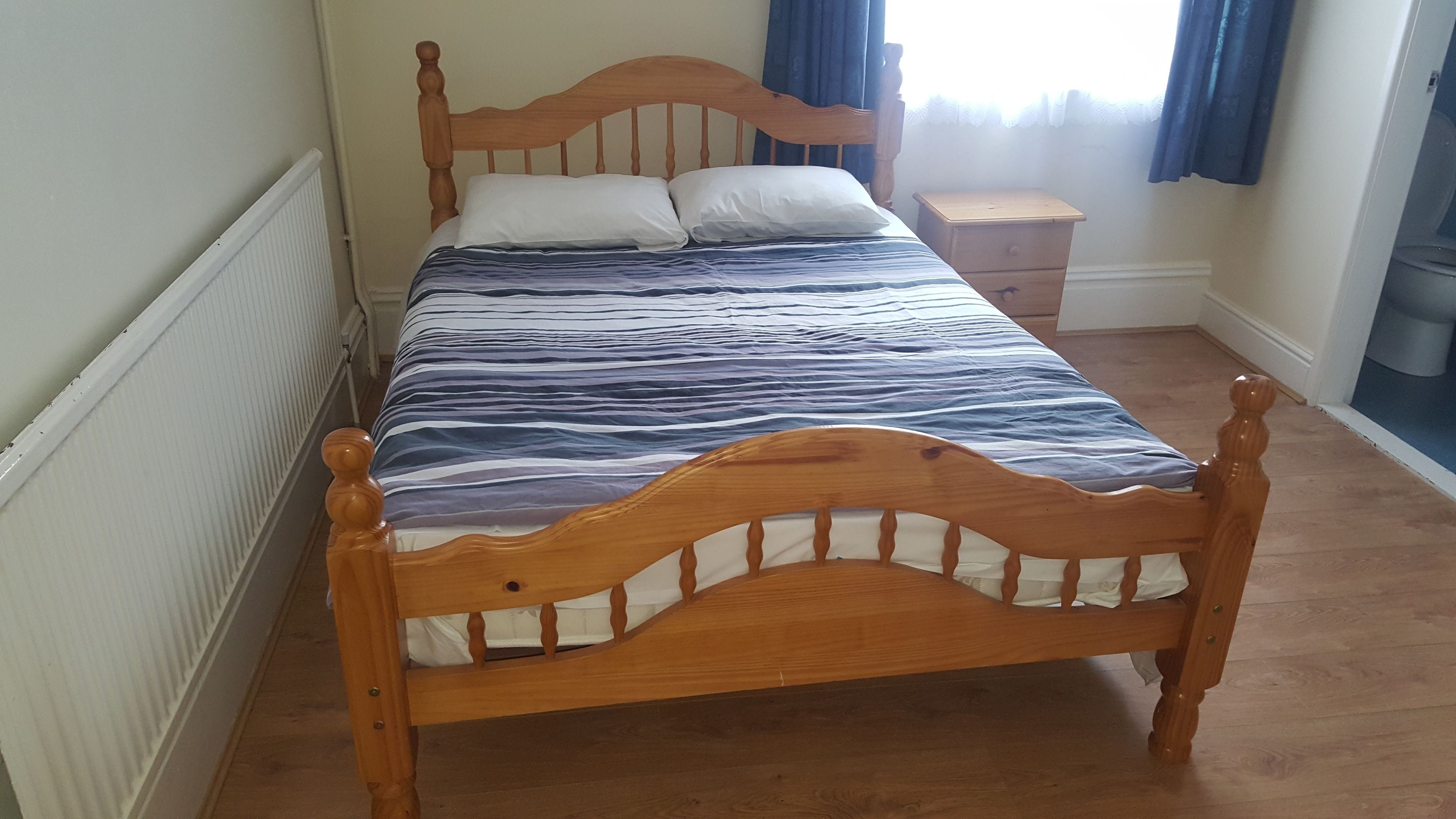 Una cama de madera con una colcha a rayas en una habitación con radiador, una mesita de noche y una ventana con cortinas.