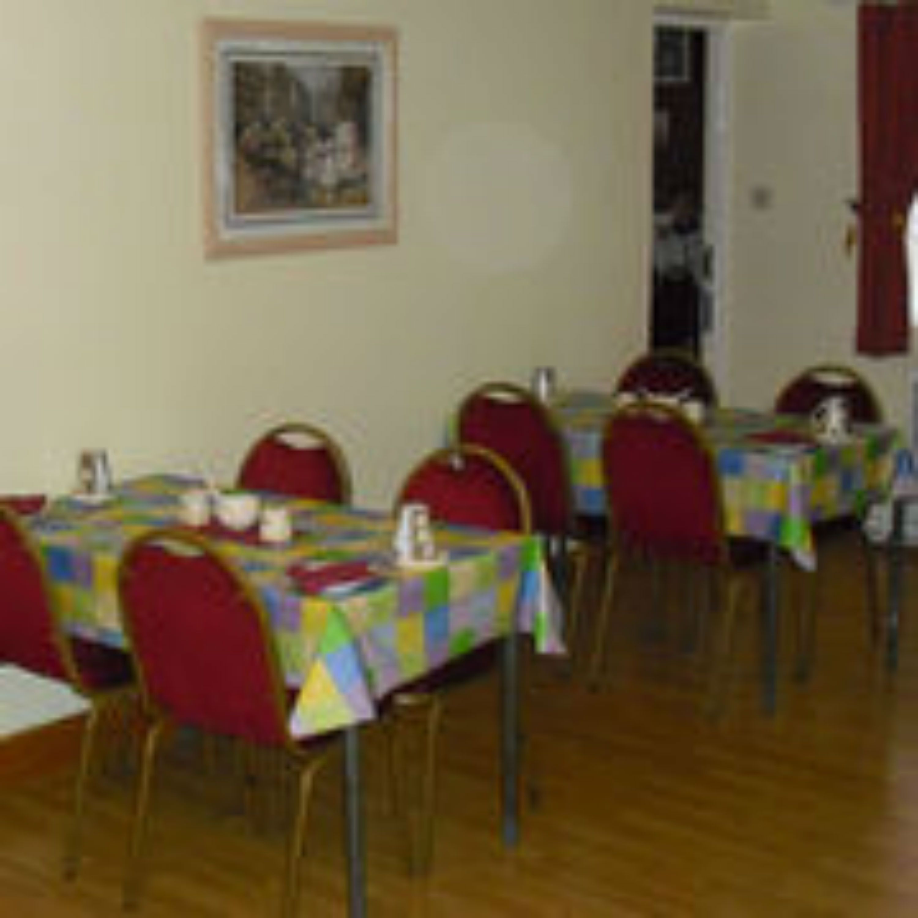 A dining area with tables covered in colorful tablecloths, red chairs, and a framed picture on the wall.
