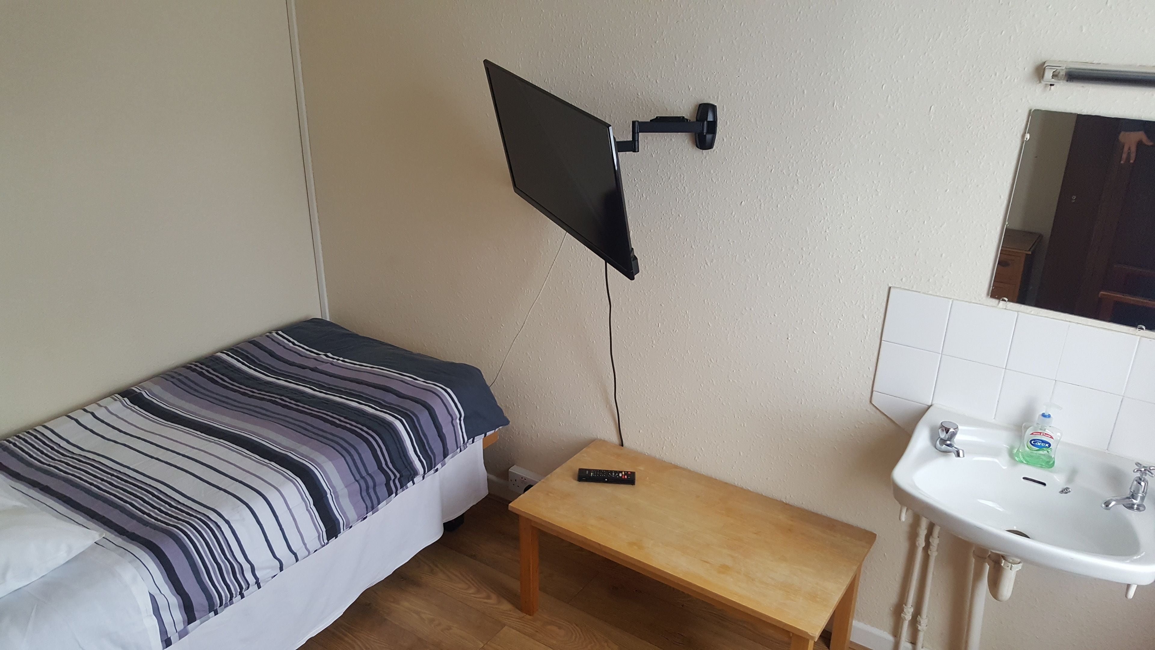 A small room with a bed, a TV mounted on the wall, a small table, and a bathroom with a sink and mirror.