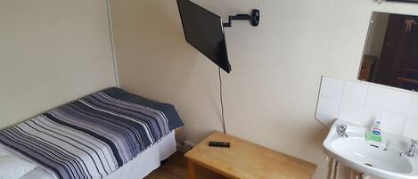 A small room with a bed, a TV mounted on the wall, a small table, and a bathroom with a sink and mirror.