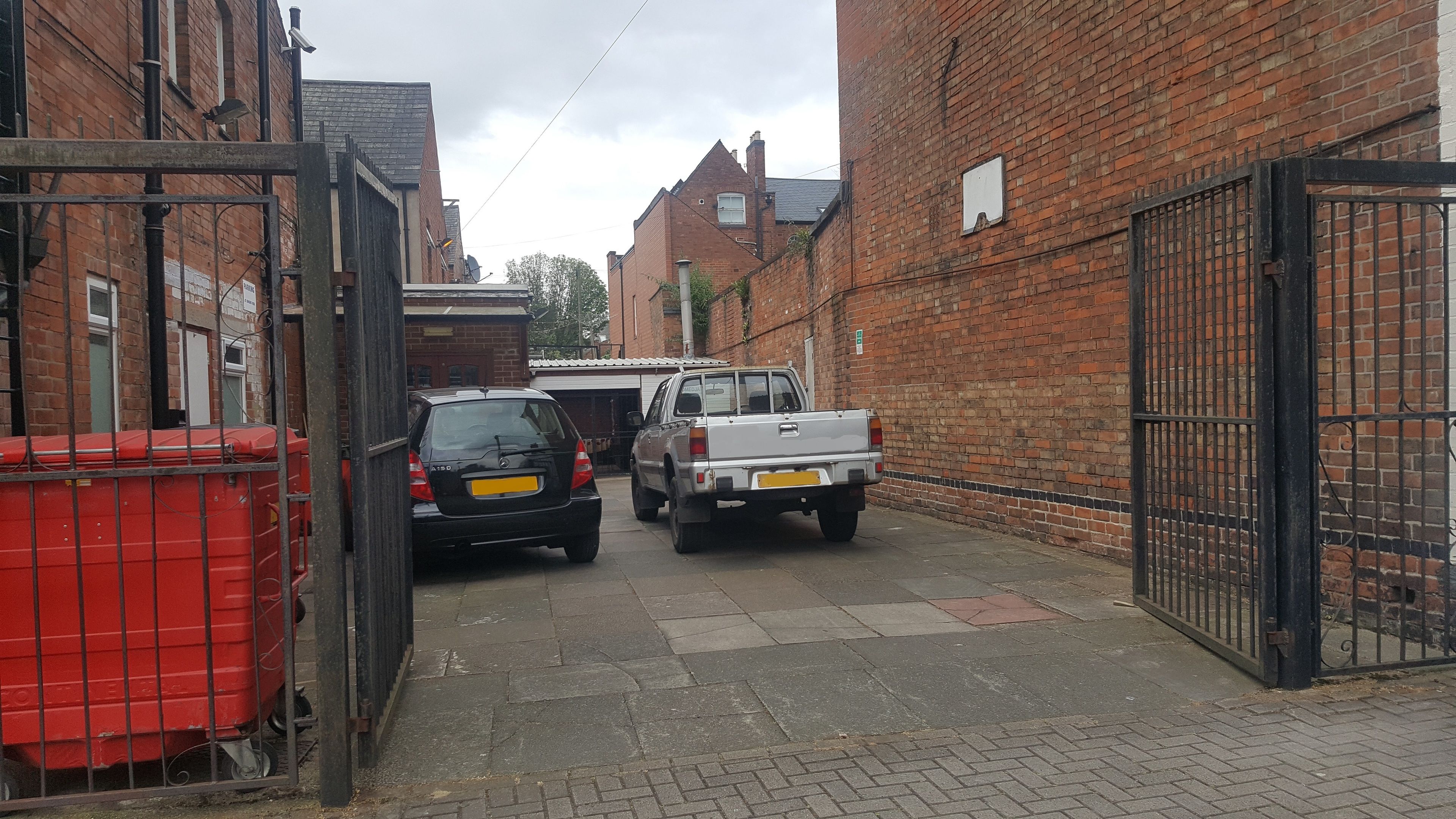 A narrow alleyway with brick buildings, a red dumpster, and parked vehicles.