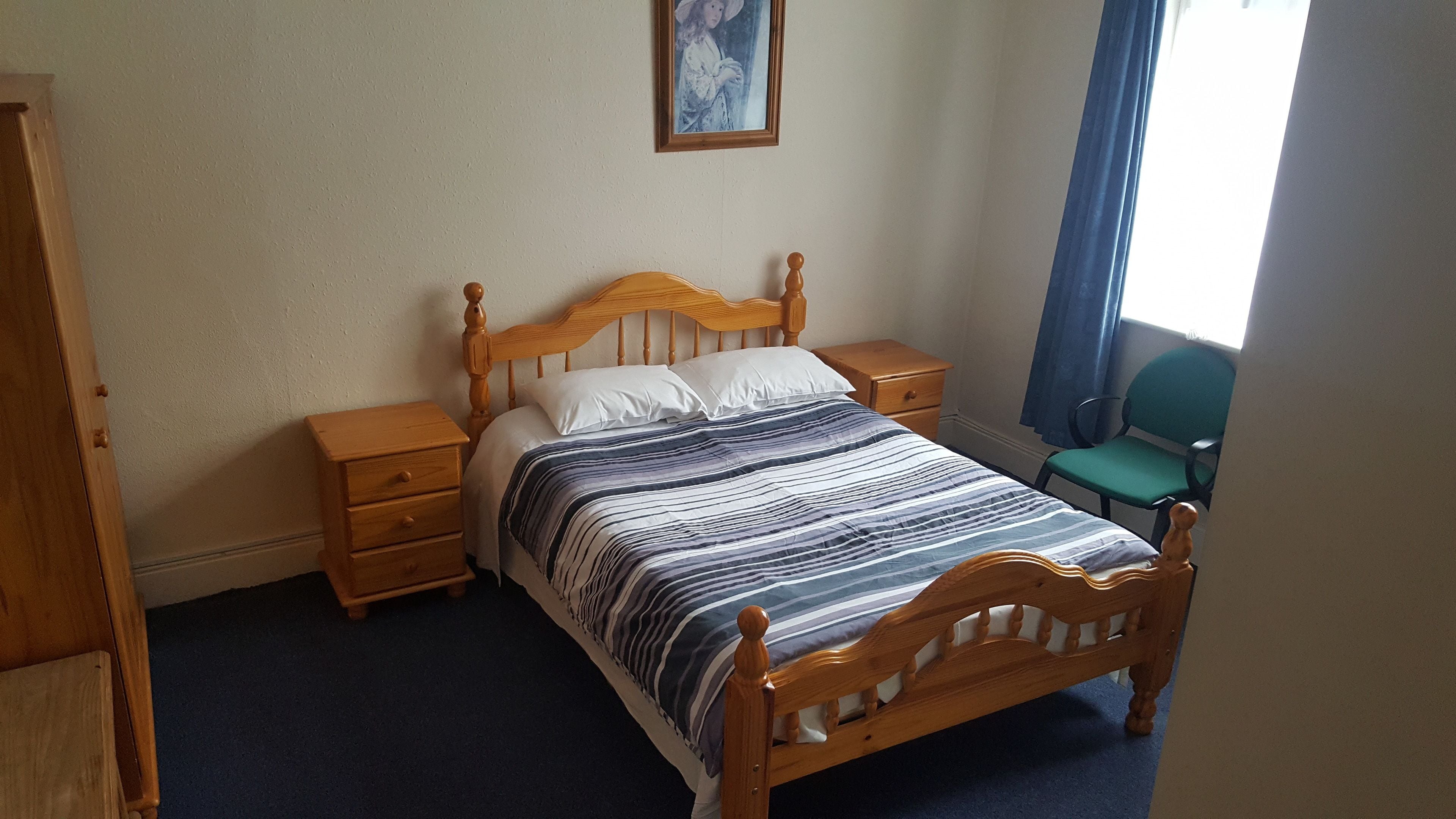 A neatly made bed with a wooden headboard, two bedside tables, a chair, and a painting on the wall.