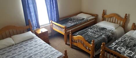 Una habitación con tres camas de madera, cada una con una colcha de diferente estampado. Hay una pequeña mesita de noche entre las camas, y una ventana con cortinas azules.