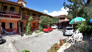 A courtyard with a red car, a white car, and a silver car parked. There are white chairs and a hammock. - Pousada Recanto de Minas (Porto Seguro)