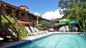 A pool area with lounge chairs and umbrellas, a staircase, and a building with a tiled roof. - Pousada Recanto de Minas (Porto Seguro)