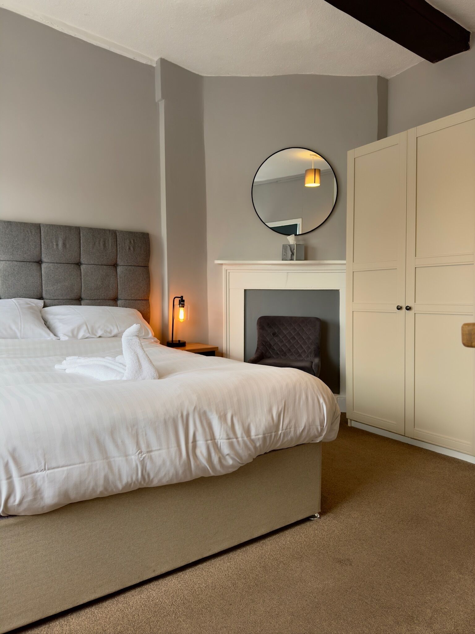 A neatly made bed with white linens, a round mirror above a fireplace, and a large wardrobe.