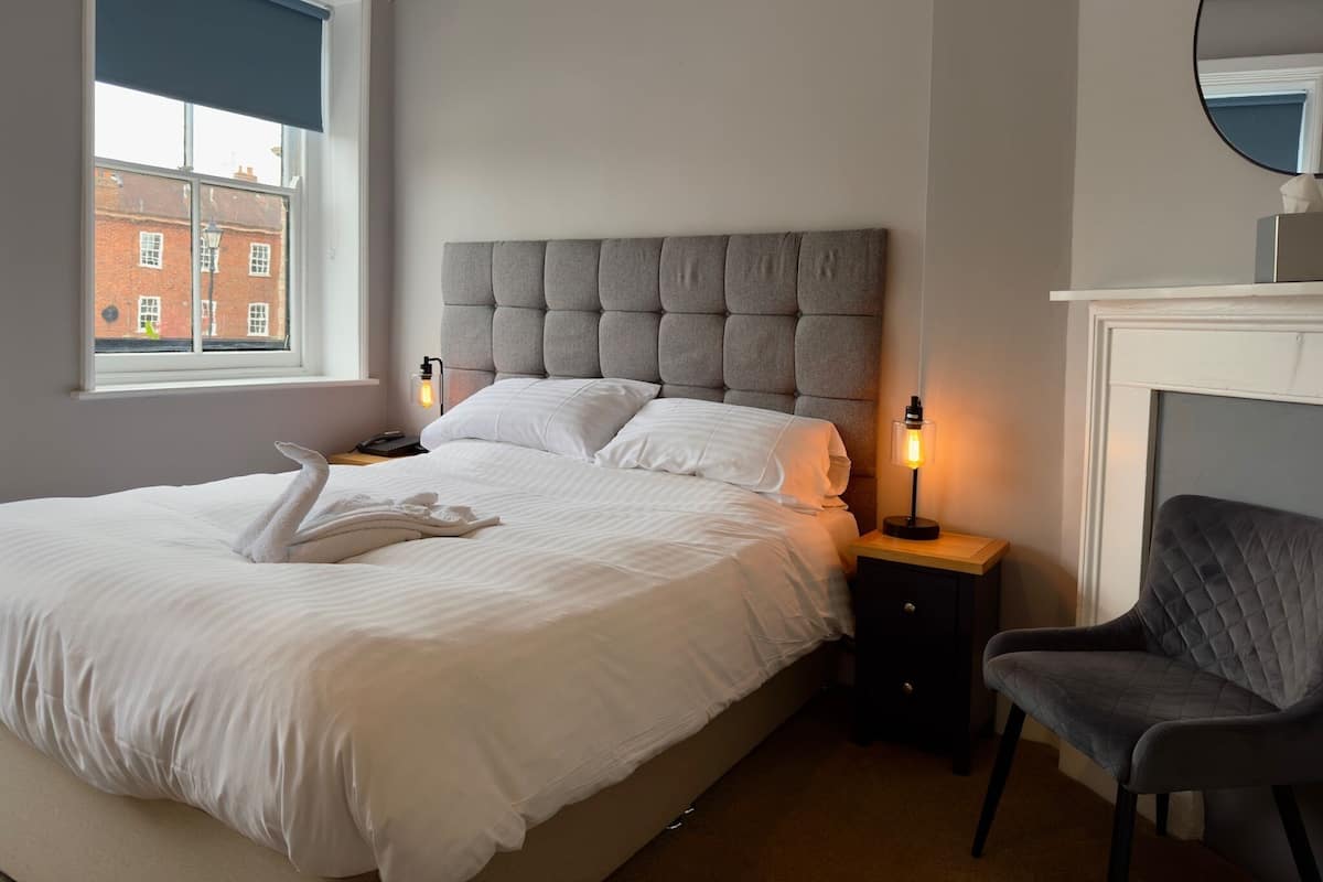 Cama bem arrumada com cabeceira estofada cinza, mesa de cabeceira com abajur, uma cadeira e vista para um edifício de tijolos através de uma janela com persiana azul.