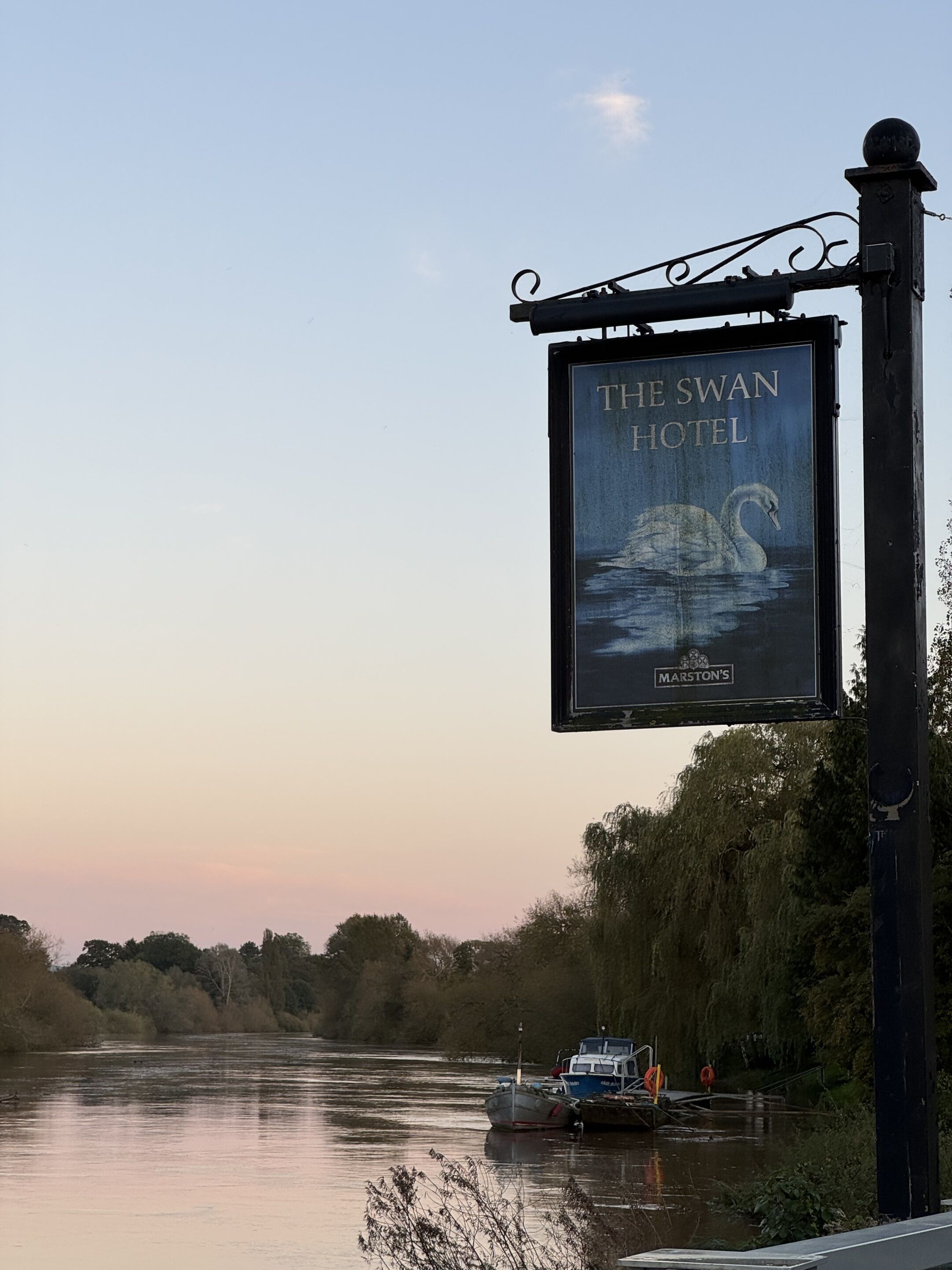 A sign for The Swan Hotel with a swan illustration, located by a river with boats.