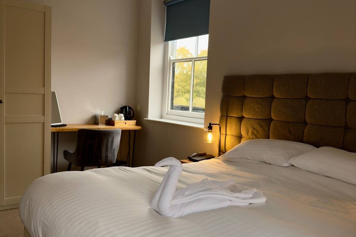 Quarto de hotel com uma cama grande, uma escrivaninha com um computador e uma janela com vista para árvores.