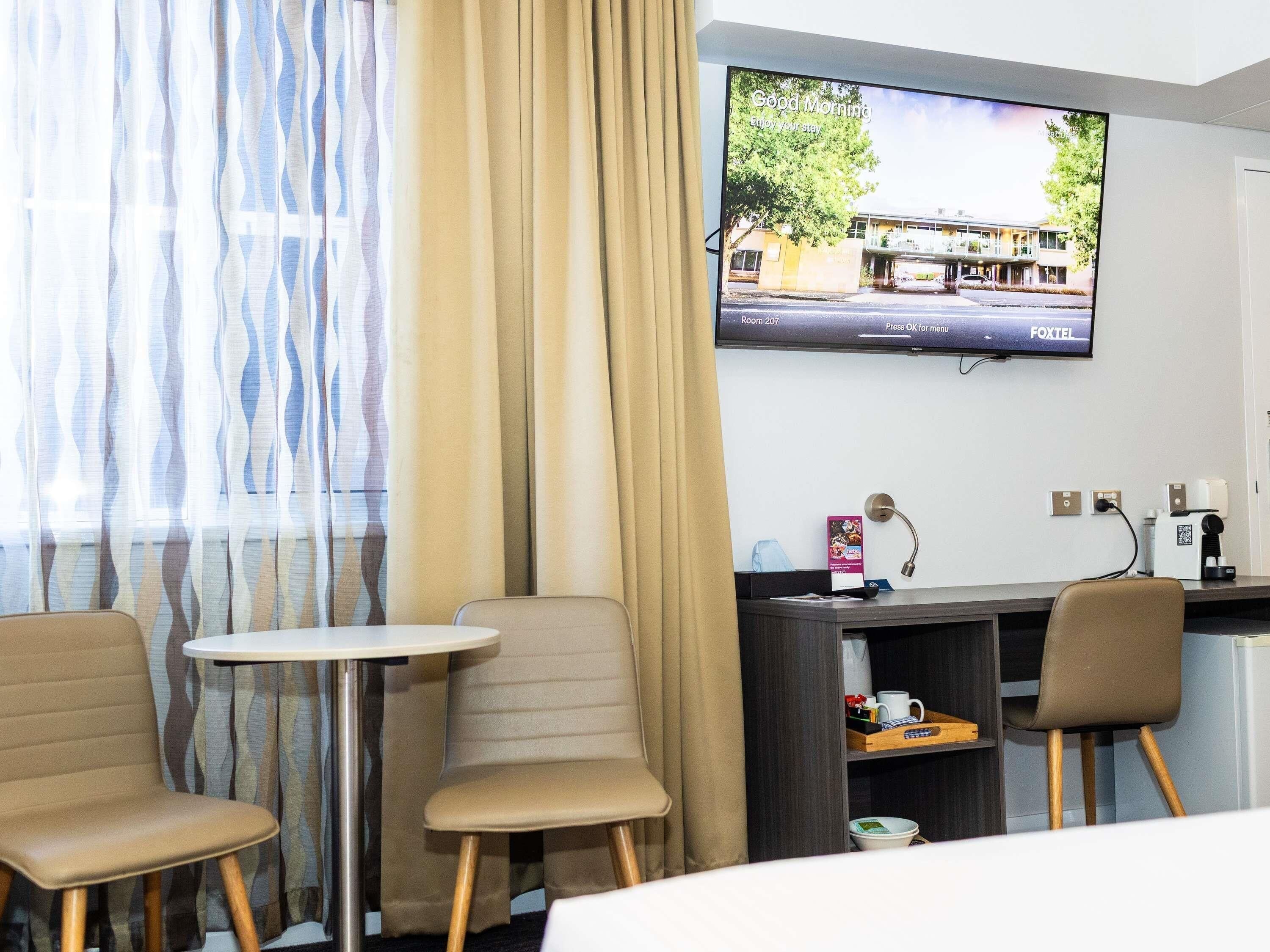 Habitación de hotel con una televisión mostrando una vista de un edificio, un escritorio con una cafetera y dos sillas.