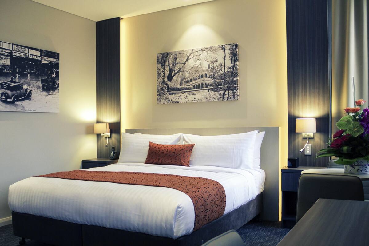 A hotel room with a large bed, two bedside tables with lamps, a wall-mounted painting, and a small table with flowers.