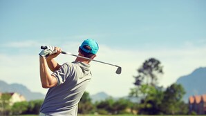 A person in a grey shirt and blue cap swinging a golf club. - Mercure Orange (Orange)